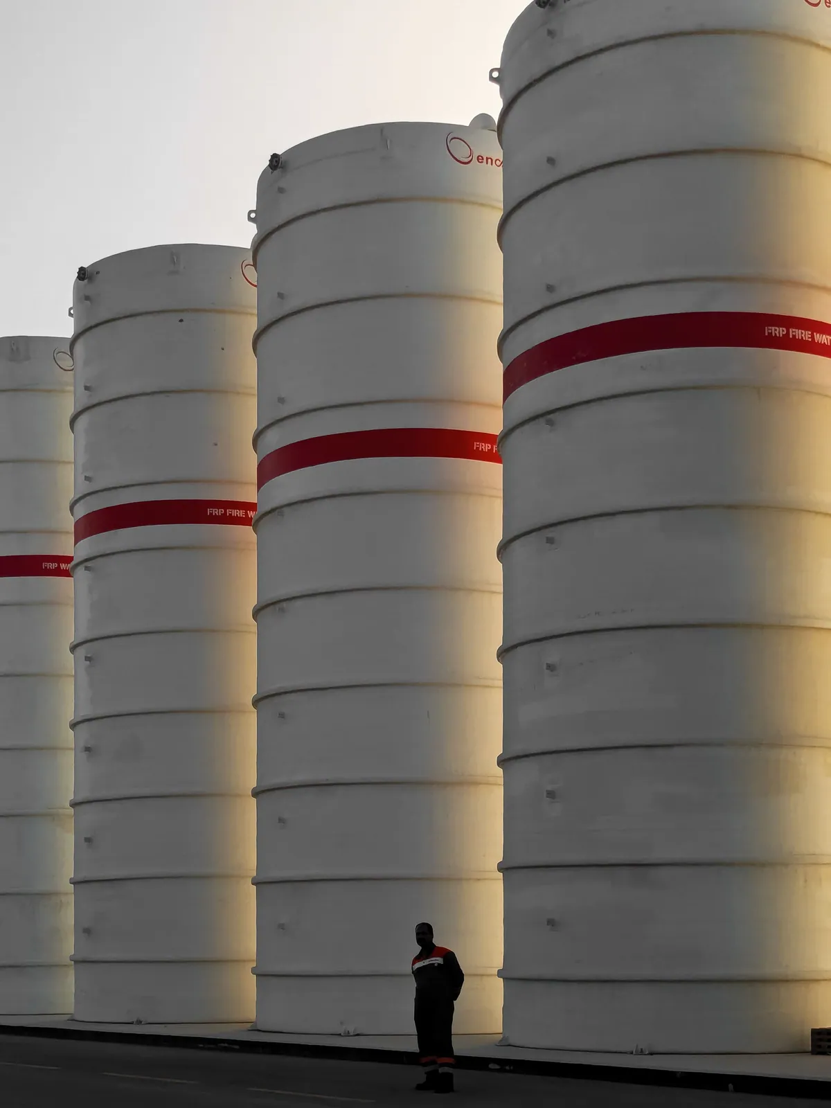 Tall steel storage tanks with red bands at sunset, a worker in the foreground for scale.